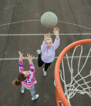 A teenage girl begins to throw a netball through a hoop whilst another girl tries to defend. They are wearing casual sports wear and the image is shot from a high angle view.